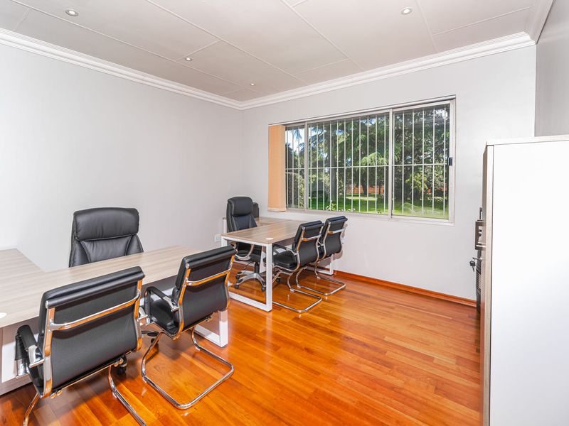 A bright office space featuring polished hardwood flooring, recessed ceiling lighting, and large windows with security bars overlooking the garden.