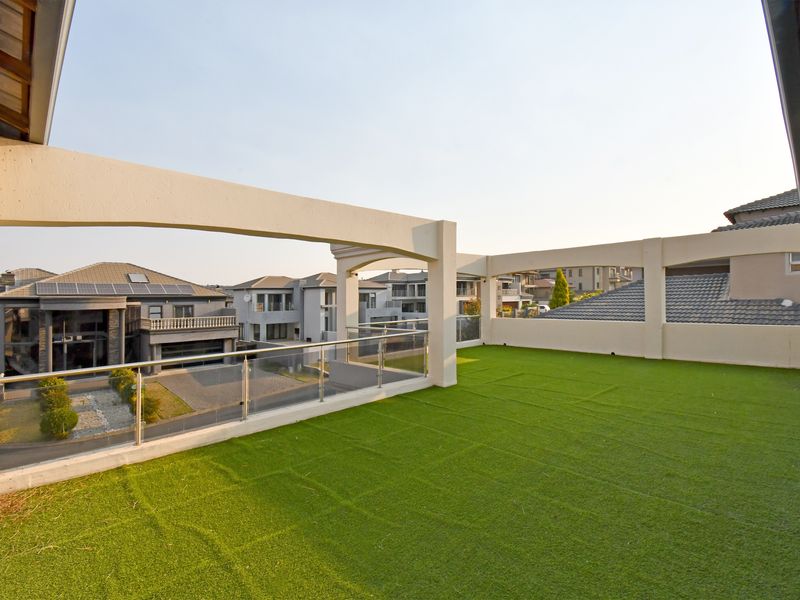 Spacious rooftop balcony featuring artificial grass, a modern stainless steel and glass balustrade, and a concrete pergola structure with views of the surrounding estate.