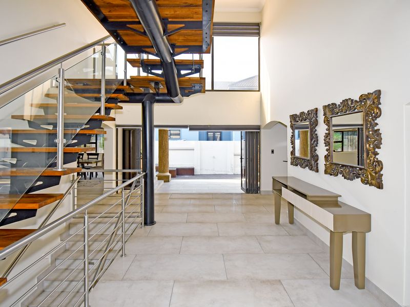 Spacious double-volume entrance hall featuring a modern open-tread staircase with glass balustrades and large format floor tiles leading to an outdoor patio area.