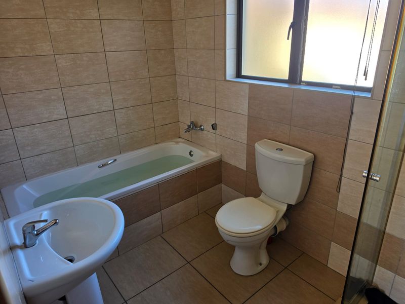 A functional bathroom featuring a bathtub, pedestal basin, and toilet, finished with neutral-toned wall and floor tiles and an obscured glass window.