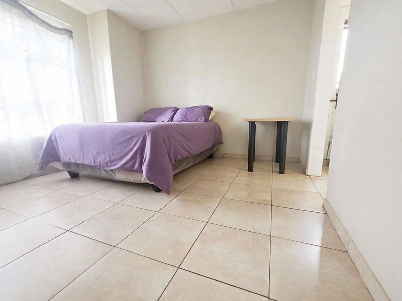 A simple bedroom featuring neutral tiled flooring and a window with security bars, offering a functional and clean living space.