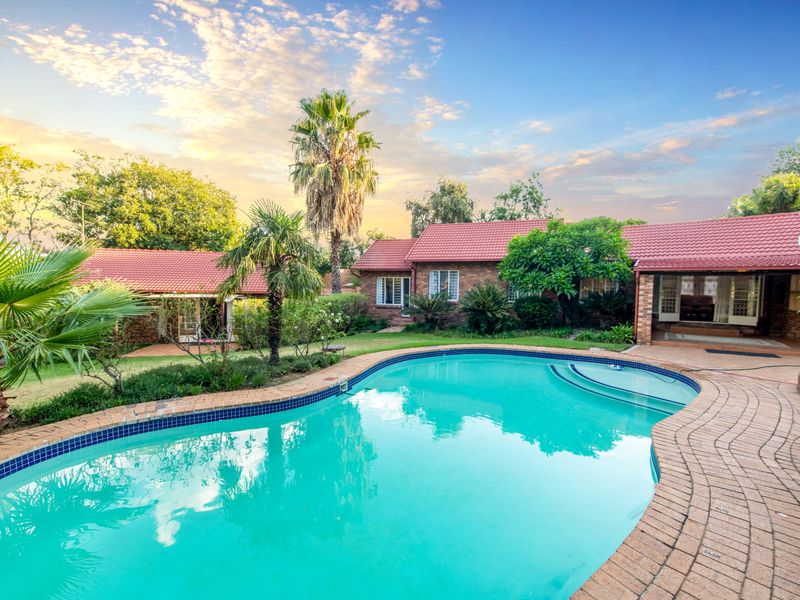 A well-maintained backyard featuring a swimming pool with brick paving, surrounded by established garden landscaping and a brick house with a tiled roof.