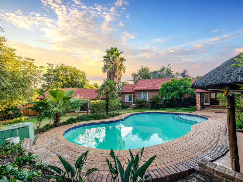 A well-maintained swimming pool area featuring a brick surround, landscaped garden, and a traditional thatched lapa, set against a scenic sunset backdrop.