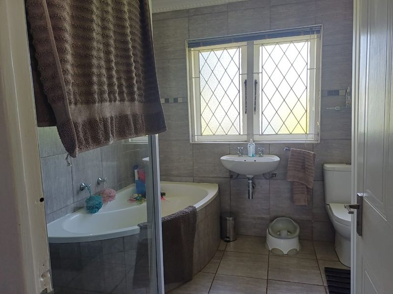 A functional bathroom featuring a built-in bathtub, wall-mounted basin, toilet, and neutral wall and floor tiling with a leaded glass window.