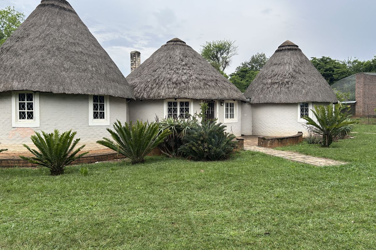 Rear view of a traditional thatched-roof dwelling featuring multiple circular structures, lattice windows, and a surrounding garden with a paved walkway.