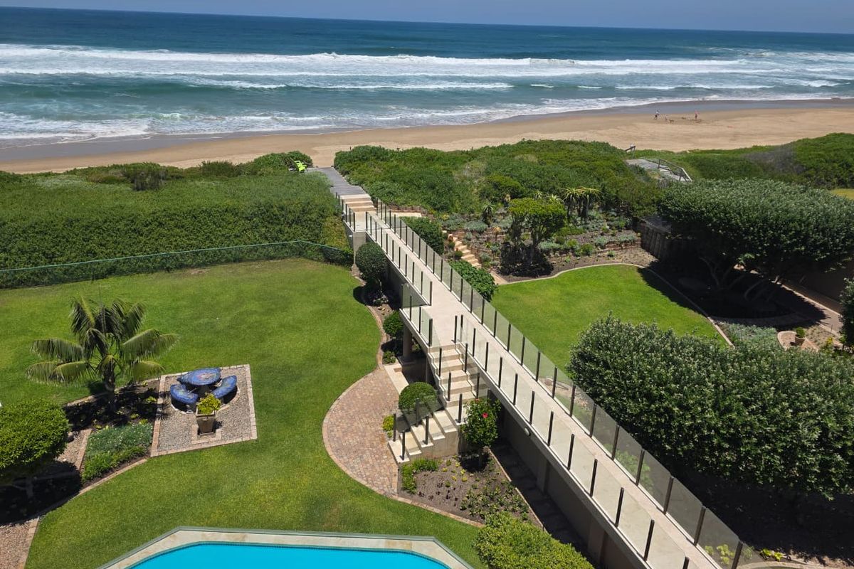 Aerial view of a coastal property featuring a swimming pool, landscaped garden, and a private elevated walkway leading directly toward the beach.