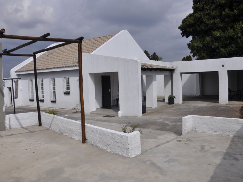 A single-story house featuring a white facade, tiled roof, and a paved driveway with an attached pergola and covered entrance area.