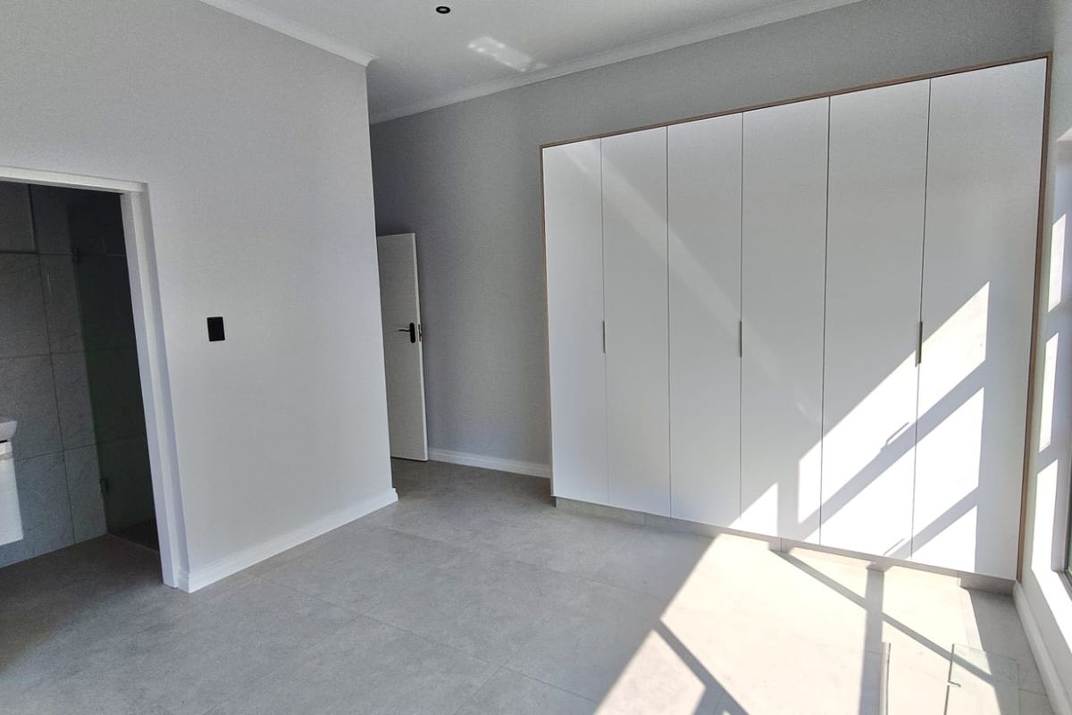 A bright, modern bedroom featuring neutral tiled flooring, ample built-in cupboard space, and direct access to an en-suite bathroom.