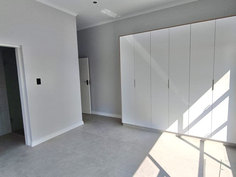 A bright, modern bedroom featuring neutral tiled flooring, ample built-in cupboard space, and direct access to an en-suite bathroom.