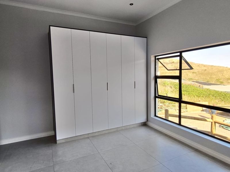 A bright, modern bedroom featuring neutral tiled flooring, built-in white cupboards, and large aluminium-framed windows with scenic views.