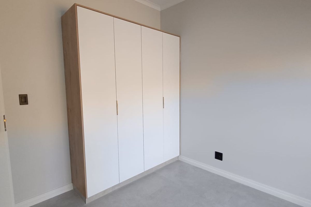 A clean, modern bedroom featuring neutral walls, tiled flooring, and contemporary built-in cupboards with a light wood-grain finish.