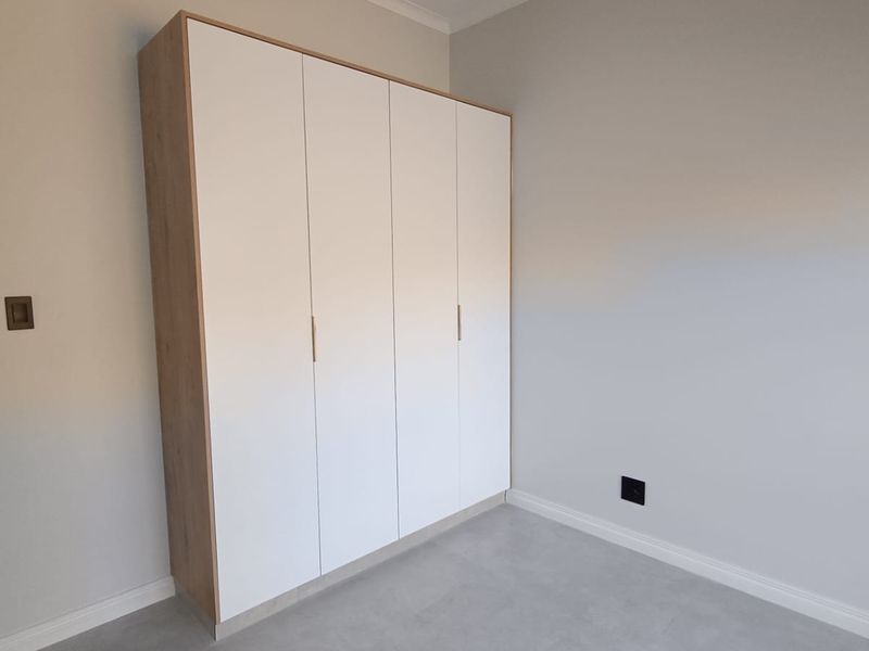 A clean, modern bedroom featuring neutral walls, tiled flooring, and contemporary built-in cupboards with a light wood-grain finish.