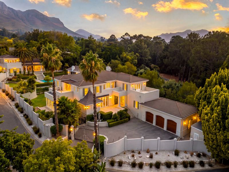 Aerial view of a luxury multi-level residence featuring a triple garage, landscaped gardens, swimming pool, and mountain backdrop at sunset.