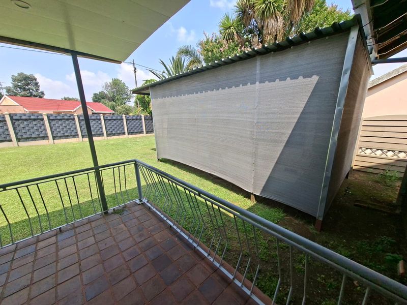 A covered patio area featuring tiled flooring and metal railings, overlooking a garden with a shade cloth structure and perimeter walling.