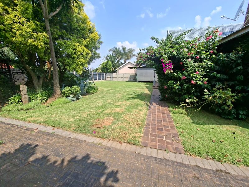 A spacious rear garden featuring a well-established lawn, mature trees, flowering shrubs, and a paved walkway leading towards the back of the property.