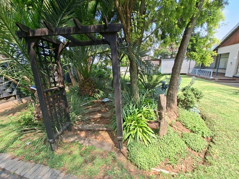 A landscaped garden area featuring a wooden pergola, mature trees, and a paved walkway leading through the greenery.