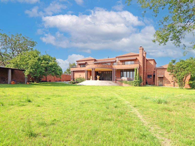 Spacious rear view of a large brick residence featuring a covered patio, upper balcony, and extensive landscaped garden grounds.
