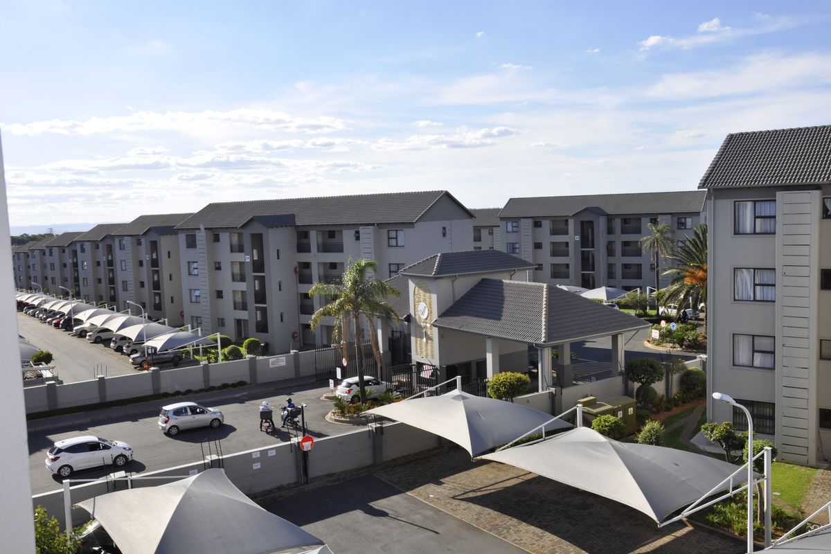 An elevated aerial view of the apartment complex, showcasing the security gatehouse, extensive shade cloth parking bays, and well-maintained building exteriors.