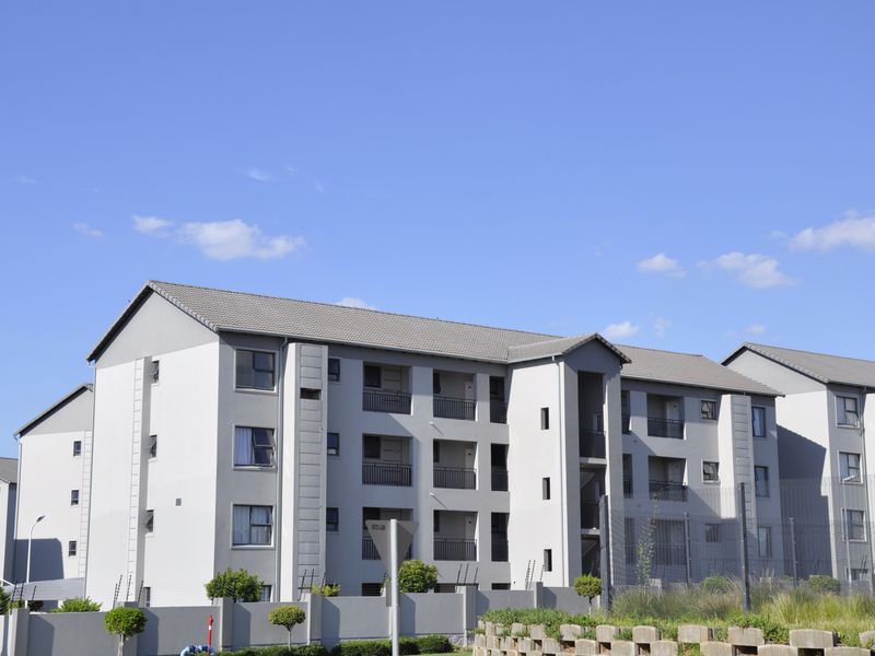 A modern multi-story apartment complex featuring a neutral grey facade, tiled roofing, and private balconies, secured by perimeter fencing and boundary walls.