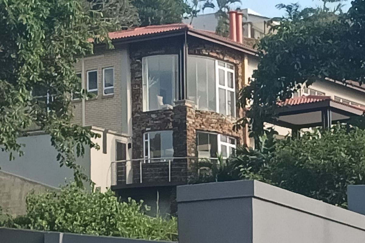 A multi-story residential property featuring stone cladding, large glass windows, and a balcony, set within a lush, elevated garden environment.