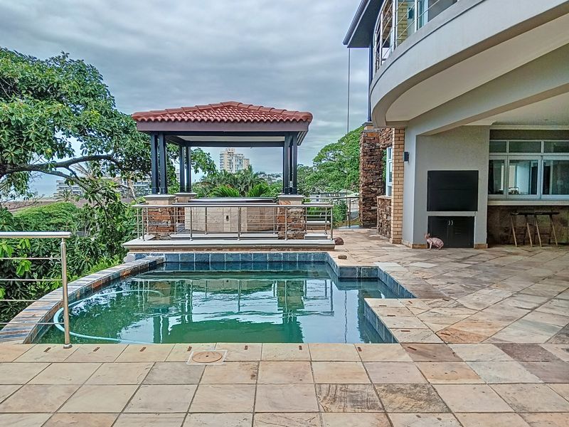 Outdoor entertainment area featuring a swimming pool, stone-paved patio, built-in braai, and a covered gazebo overlooking the surrounding landscape.
