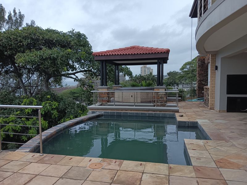 Outdoor pool area featuring a stone-paved patio, a covered gazebo, and a built-in braai, offering a functional space for outdoor entertainment.