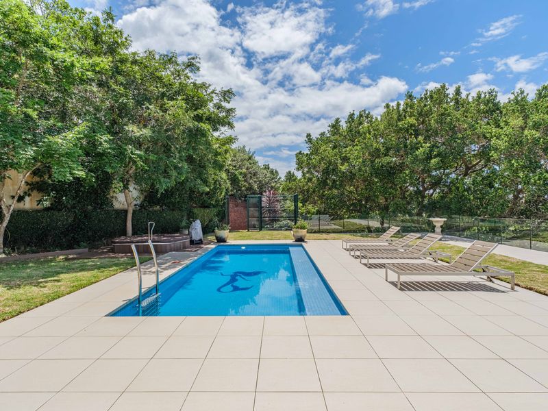 A well-maintained outdoor pool area featuring a paved deck, glass fencing, and established garden surroundings.