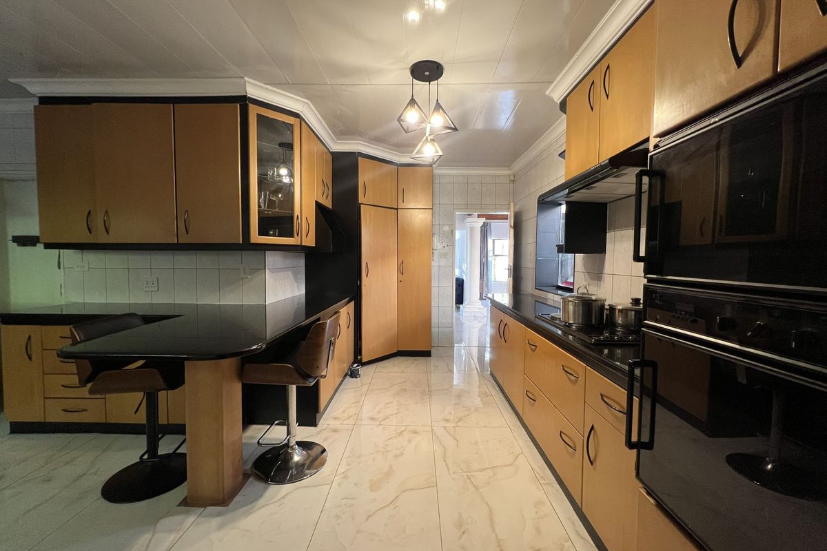 A functional kitchen featuring wood-tone cabinetry, black countertops, built-in double ovens, a hob, and polished tile flooring with a breakfast bar area.