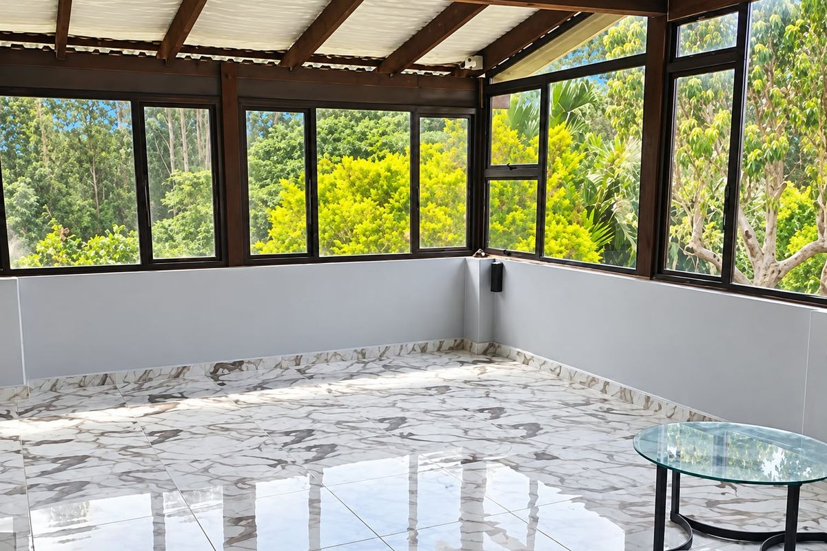 A bright, enclosed patio featuring large windows with garden views, exposed timber rafters, and polished marble-look floor tiles.