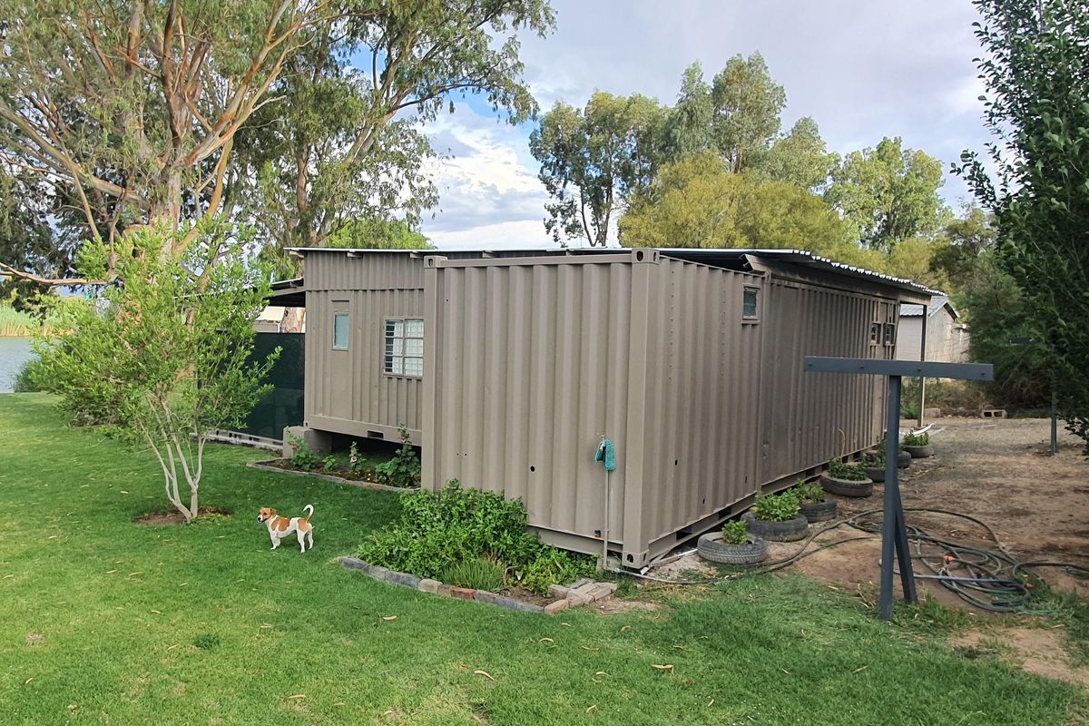 A detached container-style structure situated in a landscaped garden setting, featuring corrugated metal siding and a simple roof design.