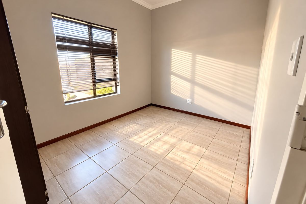 A bright, empty bedroom featuring neutral tiled flooring, clean walls, and a window fitted with venetian blinds.