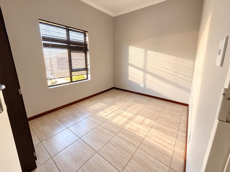 A bright, empty bedroom featuring neutral tiled flooring, clean walls, and a window fitted with venetian blinds.