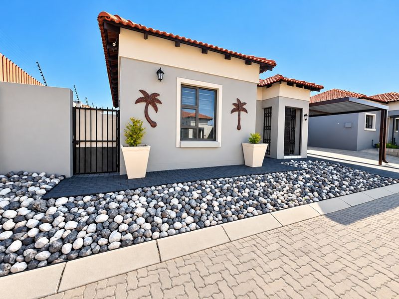 A well-maintained townhouse exterior featuring a tiled roof, paved driveway, carport, and modern pebble landscaping with secure gated access.
