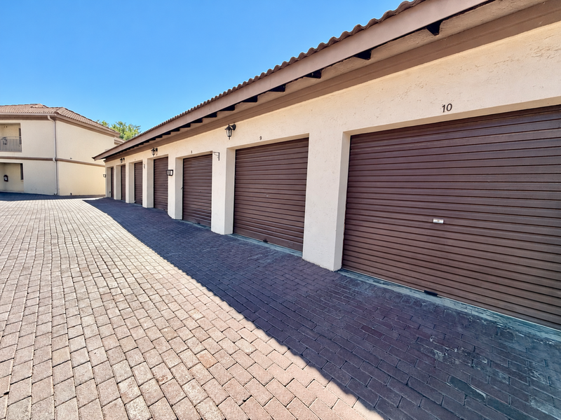 A row of secure, numbered garage units with brown roller shutter doors and a paved driveway in a well-maintained apartment complex.