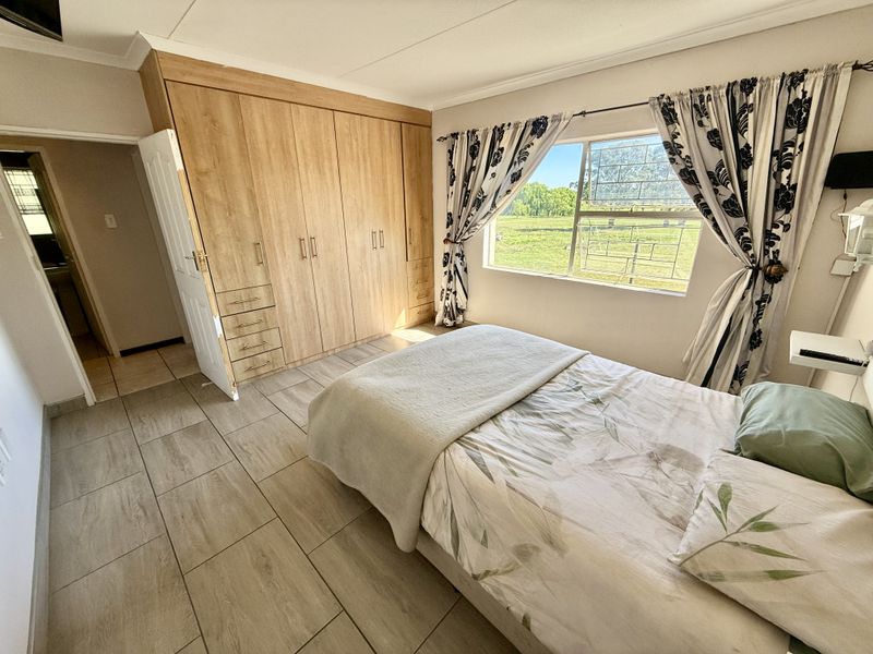 A well-lit bedroom featuring built-in wooden cupboards, tiled flooring, and a window offering a pleasant outdoor view.