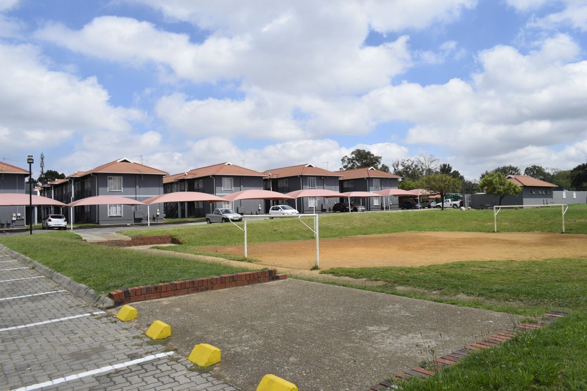Exterior view of a townhouse complex featuring communal parking with shade cloth carports and an open grass sports field area.