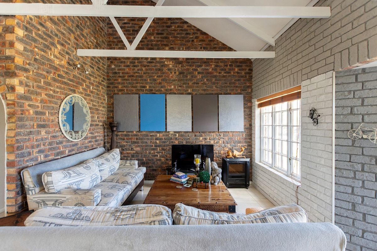 A cozy living room featuring exposed brick walls, a high vaulted ceiling with white trusses, and large multi-pane windows providing natural light.