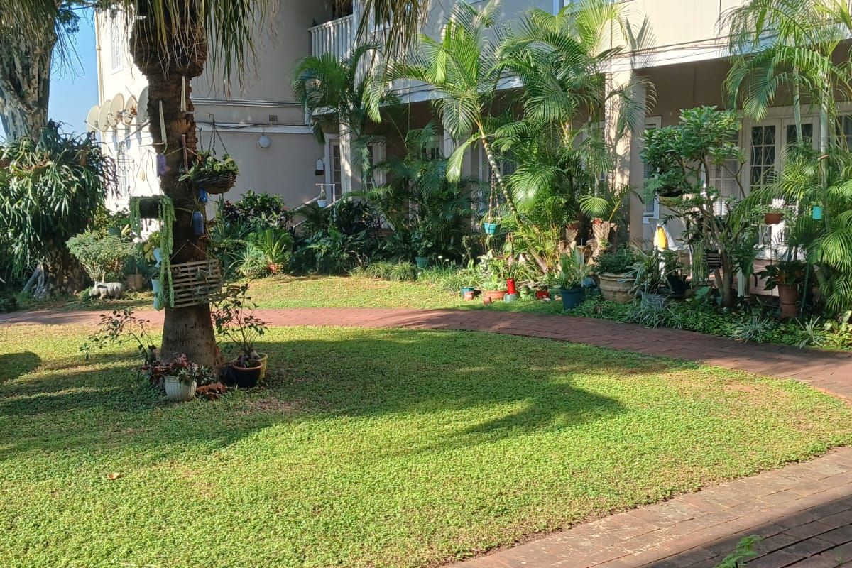 A well-maintained rear garden area featuring a brick paved walkway, lush landscaping, and access to upper-level balconies of the apartment building.