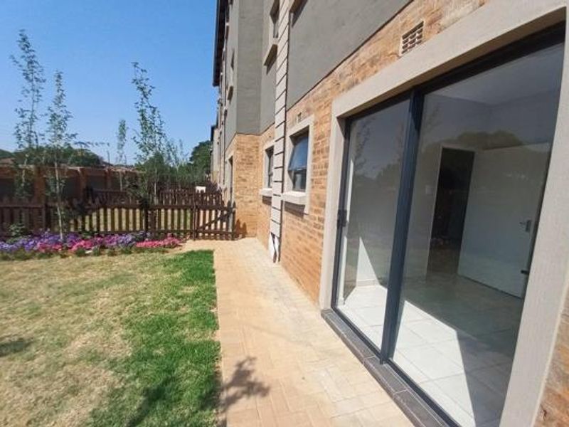 Rear view of the apartment building featuring a paved walkway, private garden area with timber fencing, and large sliding glass doors.