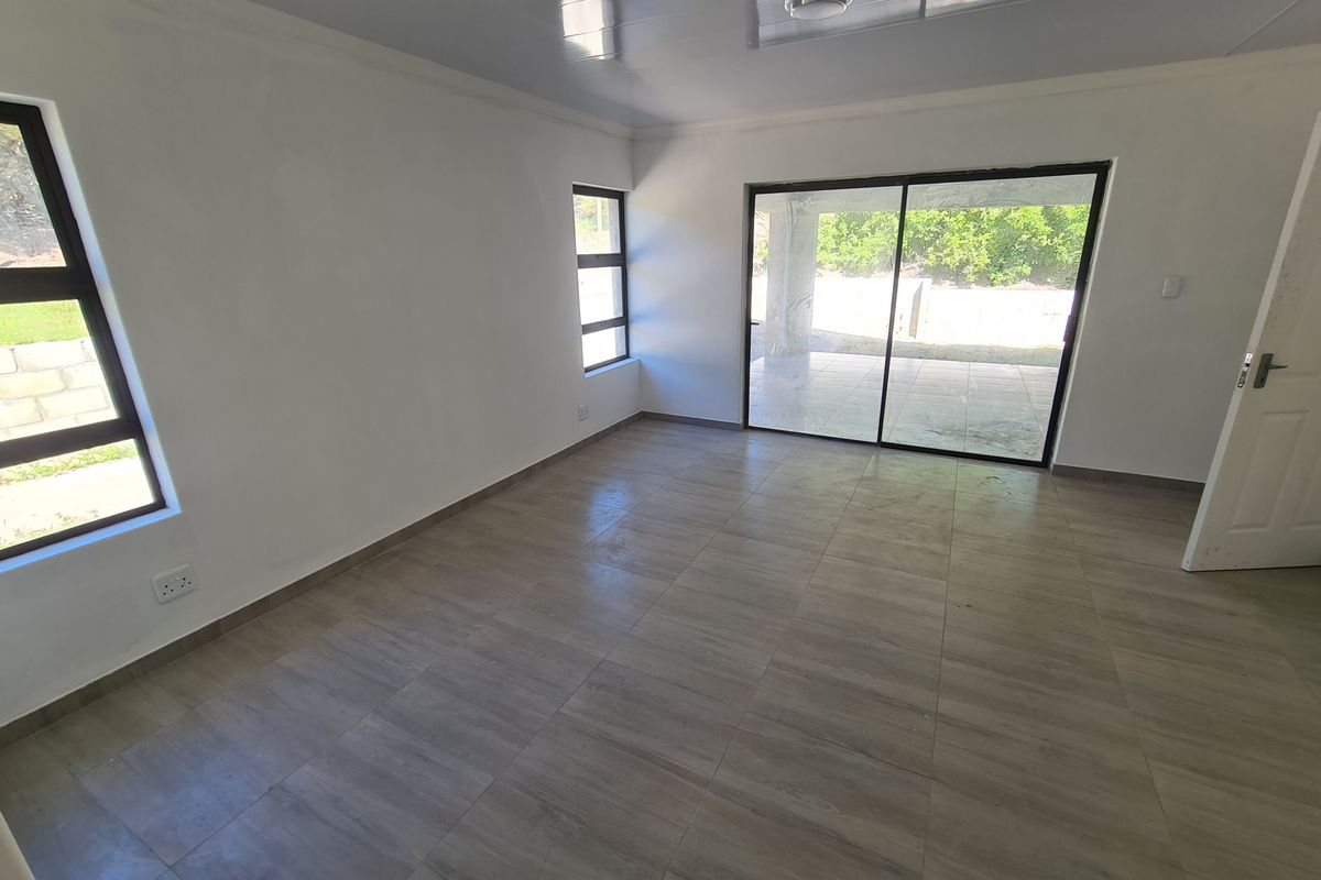 A bright, empty living room featuring modern wood-look floor tiles, large aluminium windows, and a sliding glass door leading to an outdoor patio area.
