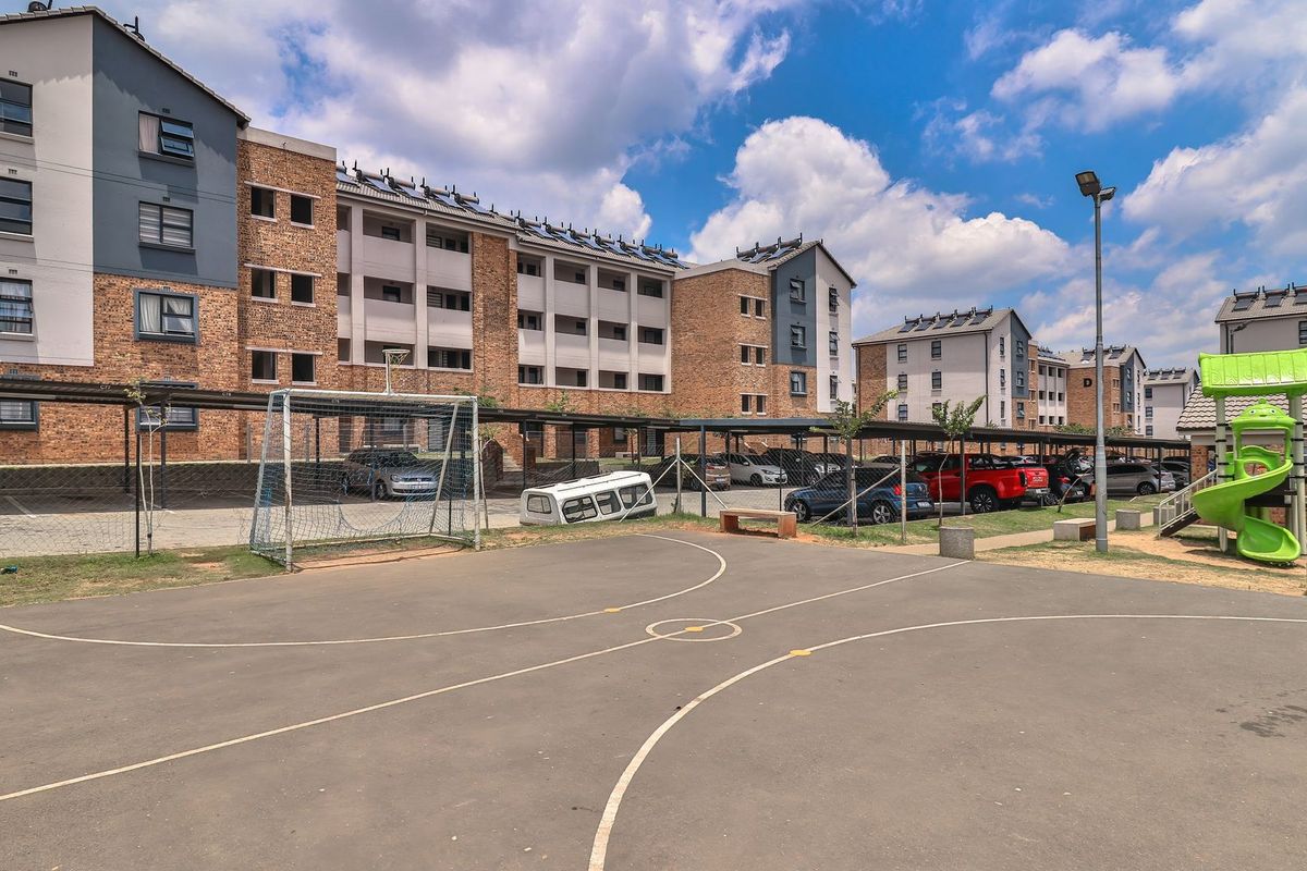 Exterior view of the apartment complex featuring a paved sports court, covered parking facilities, and communal playground area with brick building facades.