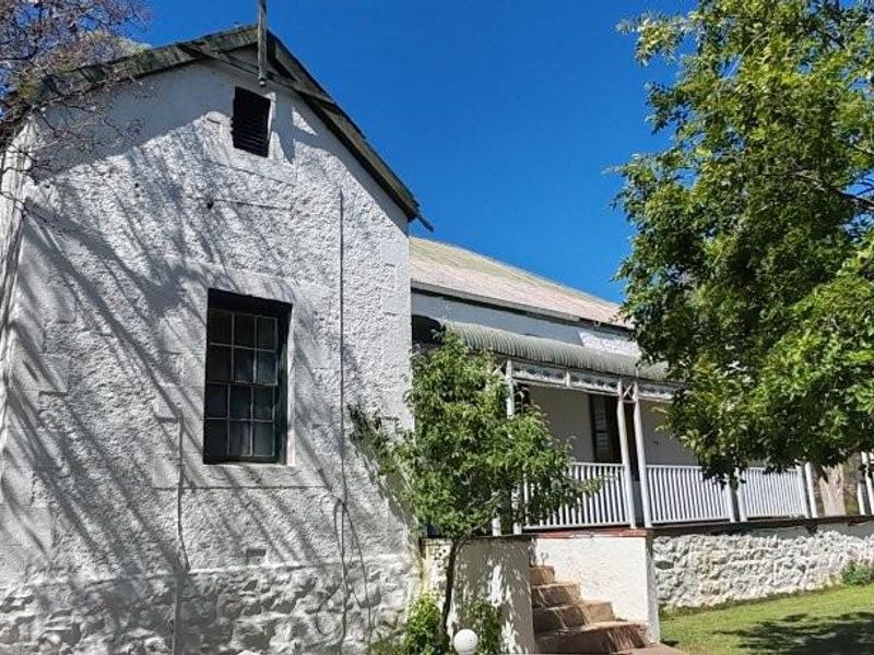 A charming farmhouse exterior featuring a classic white textured facade, a covered veranda with traditional railings, and established garden surroundings.