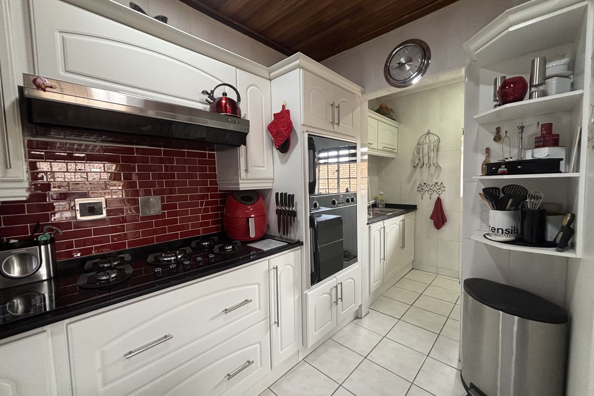 A well-maintained kitchen featuring white cabinetry, granite countertops, a striking red tiled splashback, built-in oven, gas hob, and a warm timber-paneled ceiling.