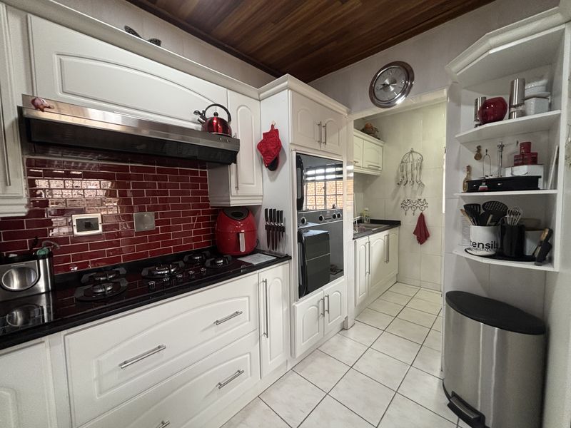 A well-maintained kitchen featuring white cabinetry, granite countertops, a striking red tiled splashback, built-in oven, gas hob, and a warm timber-paneled ceiling.