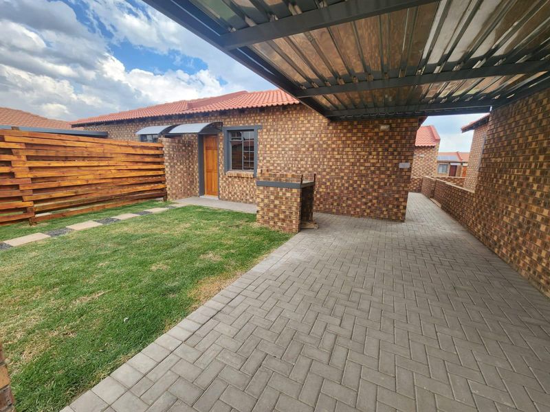 A well-maintained rear exterior featuring a paved driveway, carport, built-in braai, and a private garden area with facebrick finishes.