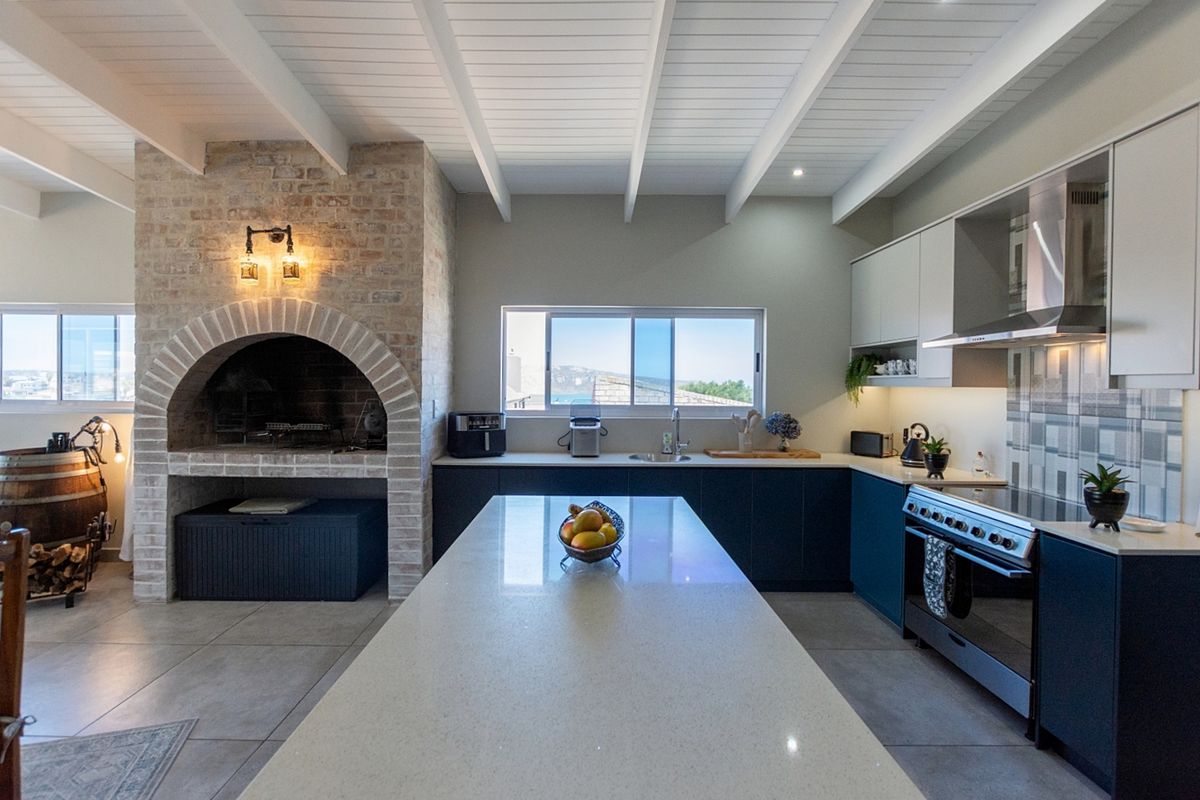 Modern kitchen featuring a large central island, quartz countertops, built-in oven, extractor fan, and a distinctive brick fireplace, all under an exposed beam ceiling.