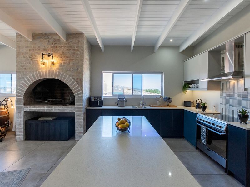 Modern kitchen featuring a large central island, quartz countertops, built-in oven, extractor fan, and a distinctive brick fireplace, all under an exposed beam ceiling.