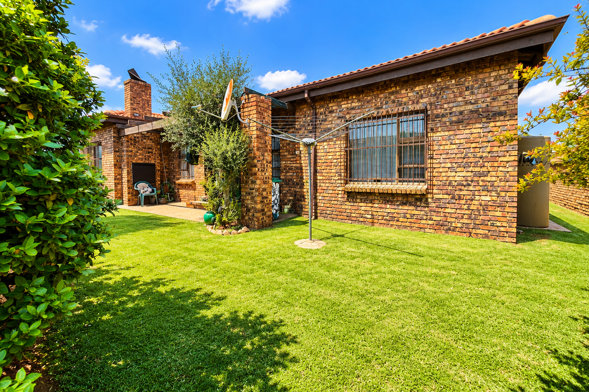 Rear view of the townhouse featuring a face brick facade, tiled roof, security bars on windows, and a well-maintained lawn with a paved patio.