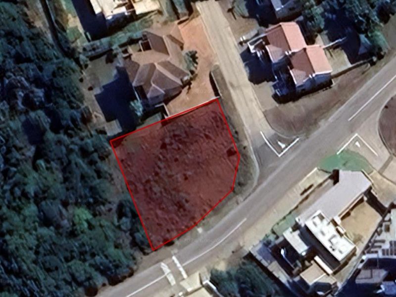 An aerial view of a vacant residential plot outlined in red, situated in a developed neighborhood with established road access and surrounding vegetation.