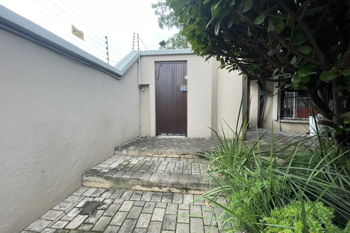 A paved rear exterior pathway leading to a secure entrance door, featuring boundary walls with electric fencing and established garden greenery.